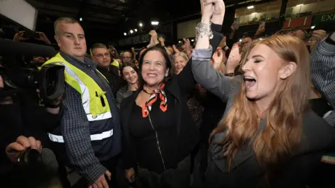 PA Media Mary Lou McDonald smiling with her arm raised in the air as she celebrates, dressed in a black top and patterened necktie... A younger woman with light brown hair, dressed in a grey blazer, is cheering and holding Mary's left hand up. To her right is a man in a checked grey shirt and hi-viz yellow waistcoat, there are press cameras visible and a large crowd of smiling supporters in the background