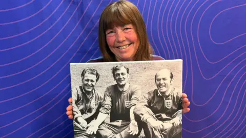 Annie Onions holds up a black and white photo of three men wearing Umbro England kit sat on a pitch with the legs crossed and their hands over knees. Dr Neil Phillips is on the right and is smiling at the camera. Onions has long brown hair with a fringe and is pictured in front of a purple background and is smiling at the camera. 