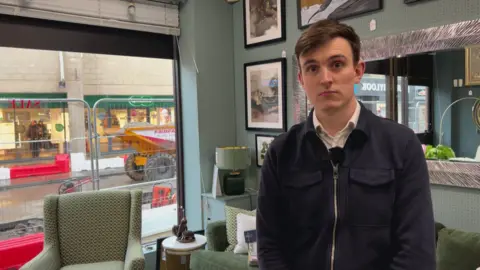 Nathan Turvey/BBC A picture of Oliver Blackburn, manager of Websters furniture store, with dark brown hair and a blue top stood in his shop, there is a chair, sofa, lamp and pictures in the background and through the shop window there are the roadworks with metal fencing.