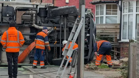 Jack Maclean/BBC An overturned lorry on its side on a residential street. A recovery team are pictured surrounding the vehicle.
