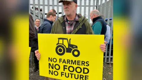 A farmer holding a yellow banner with black writing saying No Farmers, No Food, No Future and a stylised image of a tractor stands outside Hexham Market.