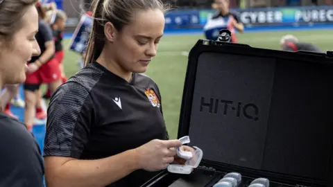 Calum Carpenter Ffion holding a white mouthguard in her hand. She stands next to a black case with multiple mouthguards in. She wears a black rugby t-shirt and stands on a rugby pitch.