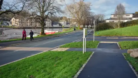 Two people walking on the new pathways either side of the road. In the background in a river, and in the foreground are the new green spaces.