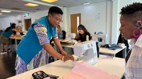 A women in a colourful dress and blue jacket reaches out to a sewing machine which another women wearing a chequered shirt is working