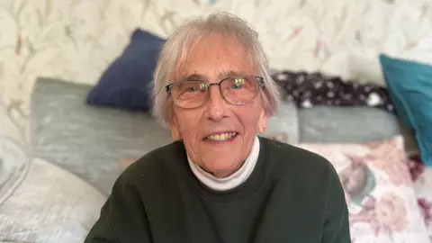 A lady with short grey hair and wearing glasses sits indoors on a sofa. She is wearing a dark green sweatshirt with ‘Coopers Hill Cheese Roll’ embroidered on it, with cushions and patterned wallpaper in the background.