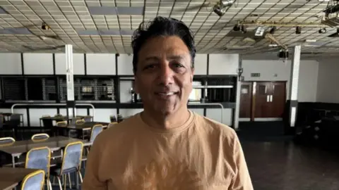 Daljit Singh inside Gurnos sports and social club wearing a yellow T-shirt with tables and chairs behind him on the left hand side of the picture.