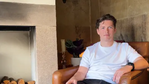 Matt Thomas with short brown hair wearing a white t-shirt and black jeans sitting in a tan coloured leather chair with a log burner to his right. He is smiling. 