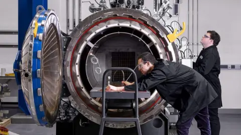 UniversityofManchester Two men in black lab coats and goggles stare into a large drum-shaped scientific machine called an autoclave. One is leaning over the machine holding equipment while the other looks on. 