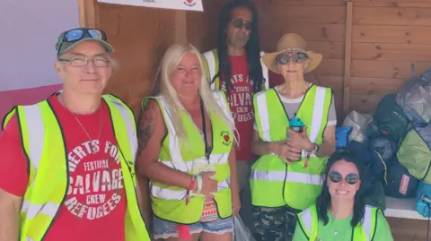 Two men and three women all wearing high vis jackets by a shed which is full of tents wrapped up in their bags. The men are wearing red T-shirts and one of the women is wearing a green T-shirt. There is a sign saying "Tent Donation Point" at the top of the shed.
