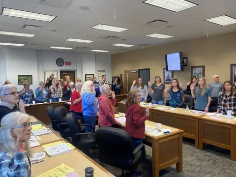 BBC / Mike Wendling A group of mostly women, casually dressed, in a community office, all raising their right hands.