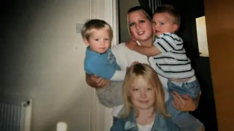 Karen Cronin A young woman with a little girl standing in front of her, holding two toddler boys in her arms.