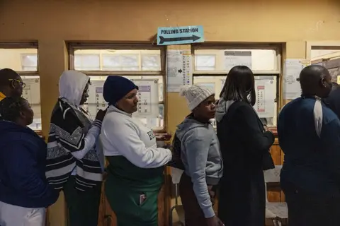 AFP via Getty Images A side view of a group of people in wearing warm clothes standing in a queue. A sign above them says 'polling station'.