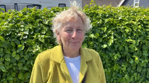 Phil Harrison / BBC A woman wearing a yellow coat and white shirt. She is stood in front of a bush with green leaves.