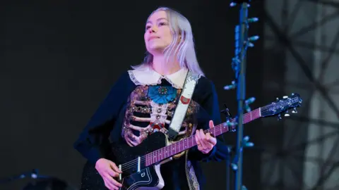 Phoebe Bridgers performs on stage holding a black guitar. She is a young woman with short, grey-coloured hair. She is wearing a black dress with a white colour and a decorated ribcage on the front of it.