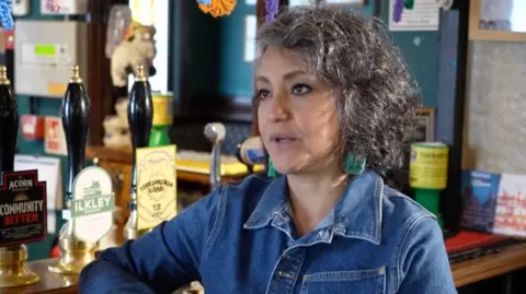 Denise stands talking to a reporter with her arm resting on the bar. She has grey hair and is wearing a denim shirt.