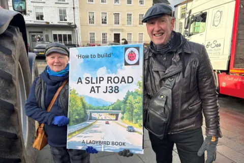 BBC/Mark McAlindon A woman wearing a blue hat, coat, scarf and gloves, dark trousers and a brown bag is standing beside a man with a grey hat, black jacket, grey gloves and dark-coloured trousers. They are both holding a placard made to look like a children's book cover. It reads: 'How to build' slip road at J38 - a Ladybird book'. They are standing between a tractor and a truck with businesses behind them.