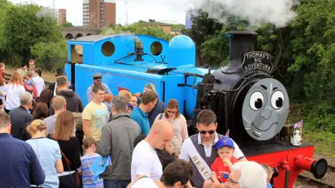 A steam engine with a blue engine cap and a black front with a black chimney billowing smoke. It has a painted face and above the face it says Thomas's big adventure. In front of the engine are men women and children, standing on a platform. 