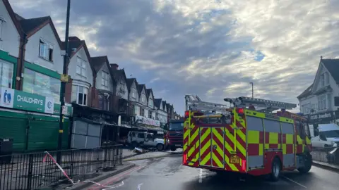 A fire engine is parked in the middle of the road near the scene of the fire. There are shops on either side of the road. There is tape on nearby railings. The burnt out shop can be seen further down from the fire engine.