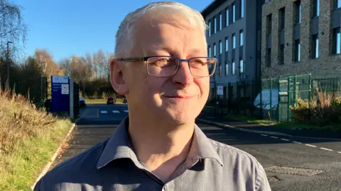Dr Robert Westgate, with short white hair, glasses, and wearing a grey shirt, looks to the right of the camera, as he stands on a road outside a large building on the right, with rectangular windows. There is a car park entrance behind him. 