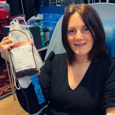 NHSBT Katie is seated in a donation chair holding up a bag of collected blood, which has a blue label reading NEO and tubing attached. The background shows medical equipment, supplies, and storage racks in a clinical or donation‑centre setting.
