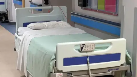 An empty hospital bed on a corridor, which has two wires coming from it. There is a small pillow and a pale green and white blanket draped over it.