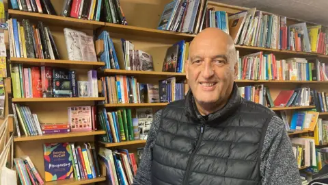 A bald middle aged man wearing a grey jumper and black gillet jacket. He is stood in front of a packed bookshelf and is smiling as he looks at the camera.