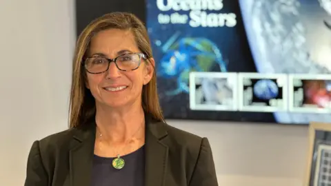 Nicole Stott, who has long brown hair and is wearing dark rimmed glasses and a dark jacket and top with a green and yellow pendant round her neck. images from stamps are on the screen behind her.