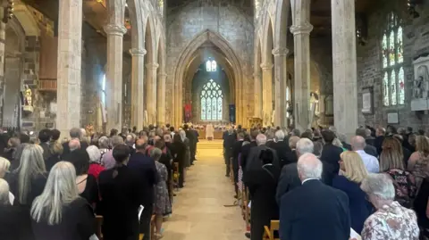 Simon Thake Hundreds of people in dark suits stand looking down a central aisle to the front of a grand Cathedral