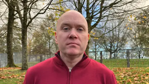 BBC A bald headed man wearing a zip up red jumper. Behind him is trees, grass and a metal fence with brown leaves on the ground.