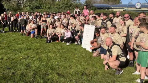 Ross Pollard/BBC A large group of people line up in identical T-shirts ahead of the Trojan Fitness memorial workout for James Kirby who was killed in Gaze