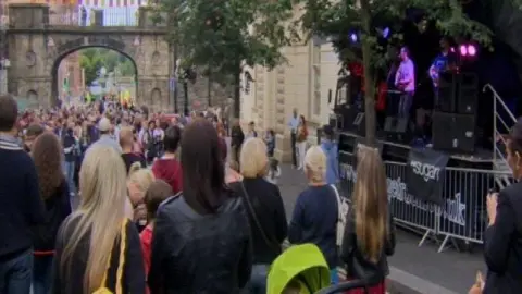 The back view of crowds gathered on a street. To the right there is a stage with a band playing. At the bottom of the street there is a stone archway.