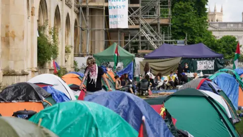 PA Media An encampment protest over the Gaza conflict on the grounds of Cambridge University. Tents can be seen outside a university building with some protest signs placed in the area.