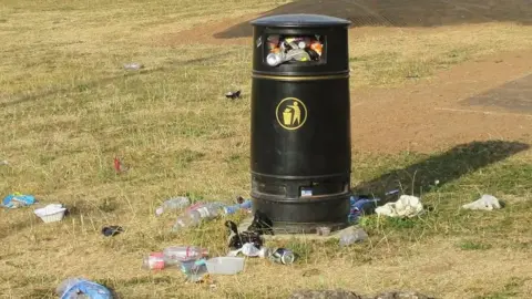 A black council park bin with a gold litter symbol on it. There is a large amount of litter on the floor around the bin and the bin itself is overflowing. 