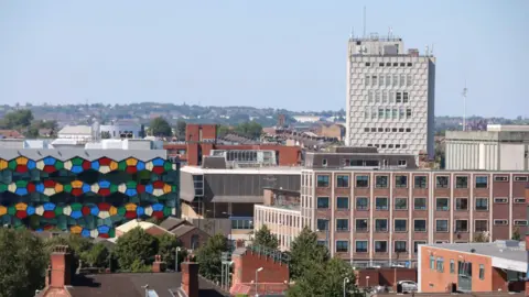 A view of a city's skyline showing a building with multi-coloured glass panels next to several big buildings, some with windows - and over them, a tall, square, cream building with windows in its centre.