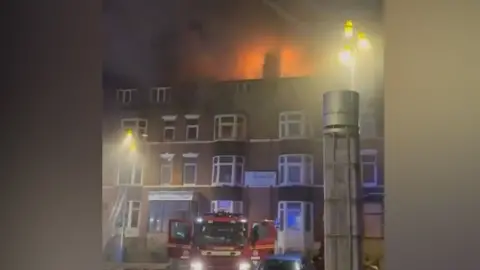 A multi-storey brick residential building at night with flames glowing along the roofline and dark smoke rising into the air. A red fire engine is parked at street level in front of the building, with blue and white emergency lights visible. Streetlights illuminate the scene, and a tall cylindrical metal structure stands to the right of the frame. Several windows are visible across the building façade, and the image appears slightly blurred, consistent with a screenshot taken in low-light conditions.