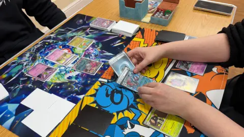 Photograph of two people playing a Pokemon card game. They're both wearing black long-sleeved jumpers, and are holding brightly-coloured playing cards with cartoon characters on them.