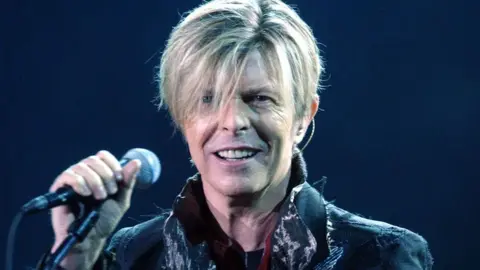 Head and shoulders of David Bowie looking at the camera while holding a microphone on stage. The background is very dark and Bowie has a long blonde fringe and is wearing a raggy-looking jacket