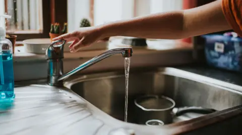A person out of vision runs a kitchen tap directly into the sink, with a saucepan sitting in the sink itself.