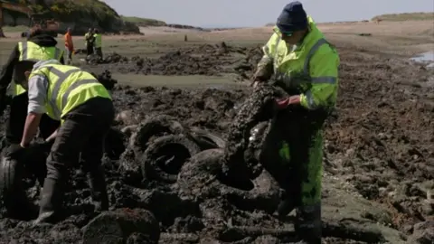 People wearing hi-vis yellow jackets, they are pulling tyres from the river mud at Ogmore.