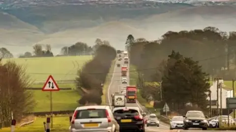 The A66 with cars travelling in either direction. The road is a single carriageway. Fields and hills can be seen in the background.