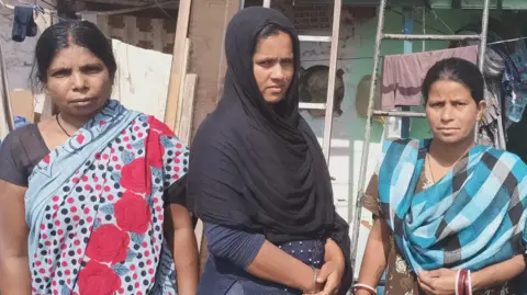Abhishek Dey/BBC Shakuntala Devi (left), wearing a white saree with floral and dotted patterns; Shakiba Bibi (centre), who has draped a black stole around her face and shoulders; and Vibha Yadav (right), wearing a turquoise and green stole over a brown kurta stand next to each other outside their residence at a slum in Delhi's Mayapuri neighbourhood on 2 April 2026. The three of them - all domestic helpers - are neighbours who have been unsuccessfully trying to buy cooking gas cylinders for three weeks. 