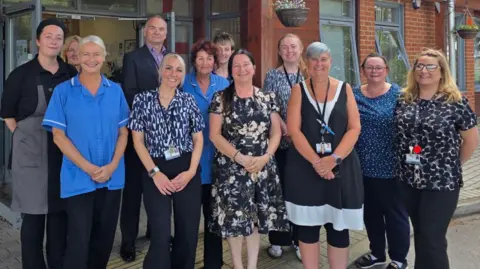 Staff members of the care home stand for a photo, they are smiling. Some wear nursing scrubs, while others wear office attire.