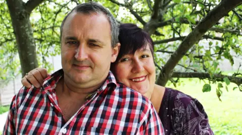 The victim Michael Ramstedt, who is wearing a black, red and white plaid collared shirt, posing for a picture beside his wife Livia, who is wearing a purple patterned blouse and resting her chin on his shoulder. They are standing under a tree in a garden, smiling at the camera.