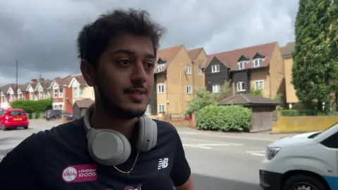 A man wearing beige headphones stood next to a main road. He is wearing a black sports top.