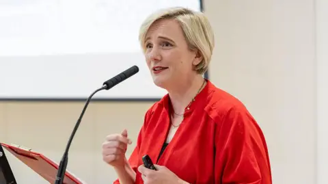 Getty Images Creasy in a bright red top speaks at a lectern while holding a presentation remote. A microphone is positioned in front of her.