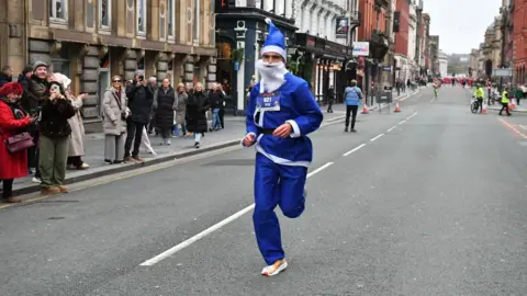 Mick Hall A man runs in a blue Santa suit on a tarmac road, with crowds watching from pavement and taking pictures. Old buildings are on both sides.