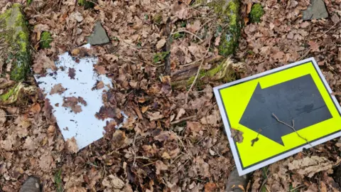 ACORN TRAILS A composite image of a race sign on the ground in Castlemilk Woods. On the left, the white back of the sign is visible underneath a pile of brown leaves. On the right, it is turned the correct way round and has a black arrow on a luminous yellow background.