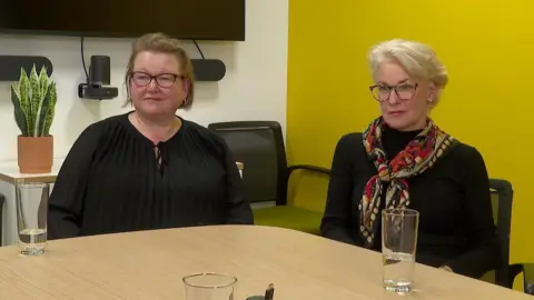 Hannah Pugliese sits on a chair in front of a wood effect table which has two water glasses on it on the left of the frame while Penny Smith sits on the right. Both are looking at the camera, wearning black tops and glasses, Penny has a colourful scarf around her neck. Behind is one white wall and one yellow wall, a snake tongue plant in a terracotta pot and the bottom of a TV screen and speakers.