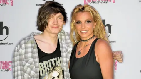 Kurt  Kurt standing with Britney, with his arm behind her. Kurtis is wearing a checked white, grey and green shirt, open buttoned, with a top underneath which has Britney spelt in green and an animated image of her on the top. Britney is smiling wearing a black vest top and white trousers, with a black neck choker and a necklace. The background is of a white advert board with promotional branding.