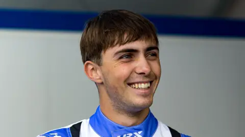 A man, Edward Pearson, in a blue and white racing suit. He is smiling and looking away from the camera.